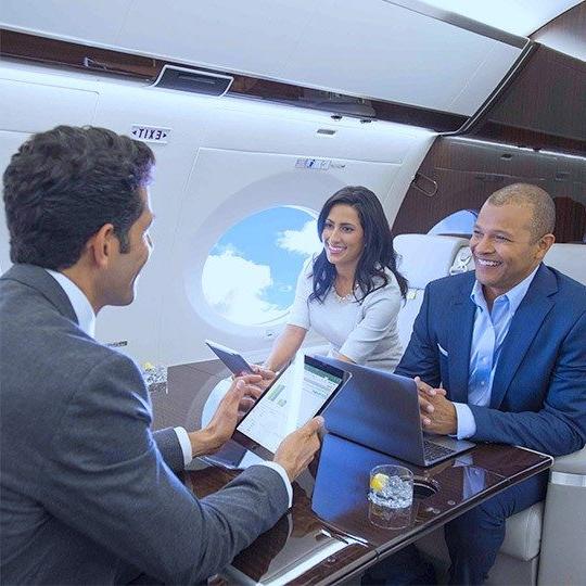 两男一女坐在一架私人飞机的桌子旁, 在私人飞机上使用平板电脑和连接WiFi的笔记本电脑