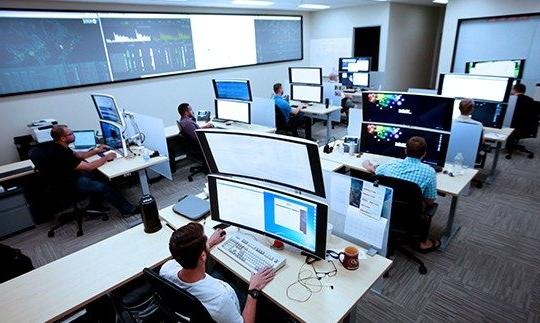 七个人坐在办公桌前，用大型电脑显示器分析数据